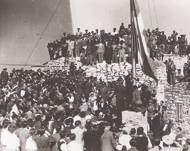 18 Oκτωβρίου 1944. H μεγάλη στιγμή. O πρωθυπουργός Γεώργιος Παπανδρέου υψώνει τη σημαία. O ελληνικός λαός μπορεί και πάλι να αρθρώνει χωρίς φόβο τη λέξη ελευθερία.