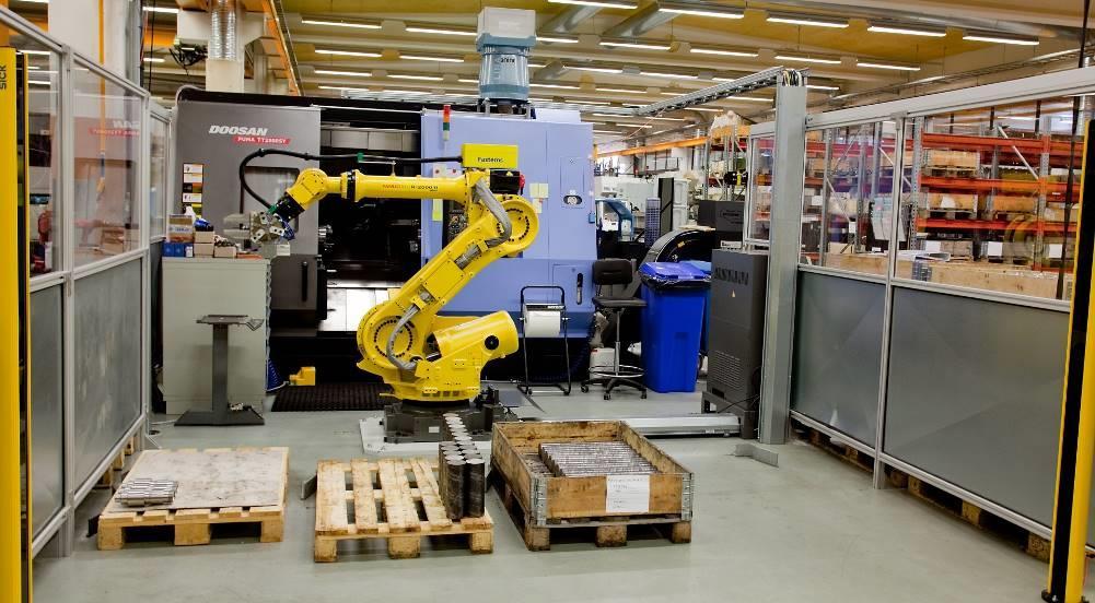 pavaizduotos tekinimo staklės su joms pritaikyta automatizacijos sistema, kurią sudaro robotas, valdymo sistema, apsauginės sienelės ir apsauginiai vartai.
